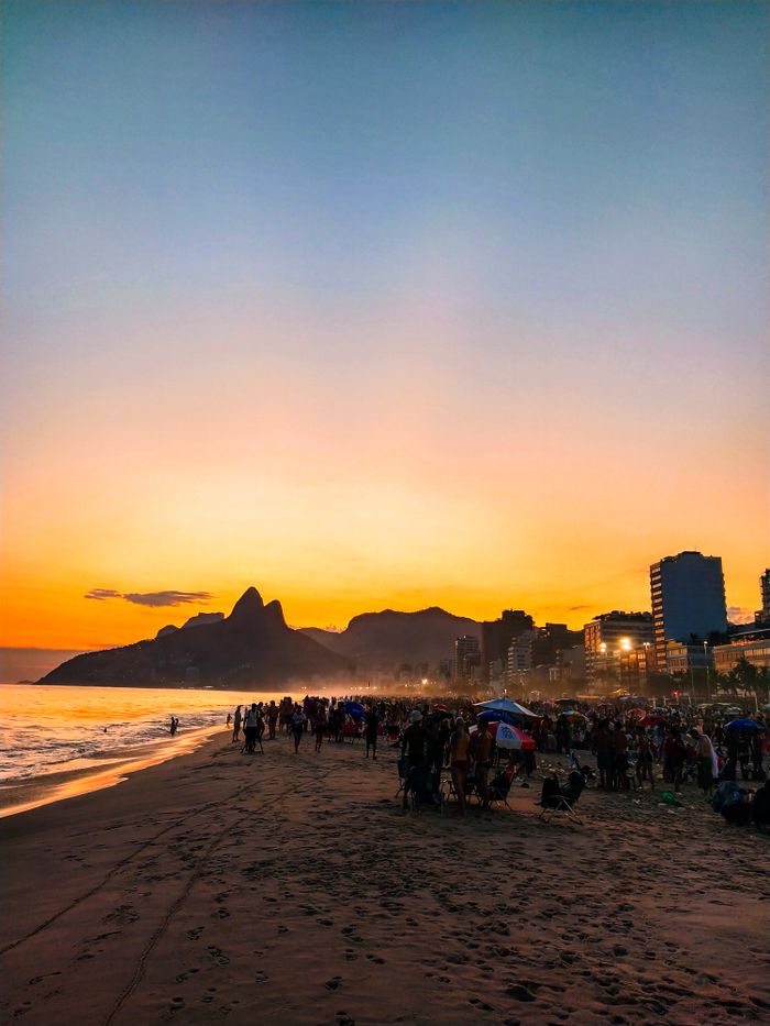 A vibrant sunset over Ipanema beach filled with people and a breathtaking view of distant mountains.