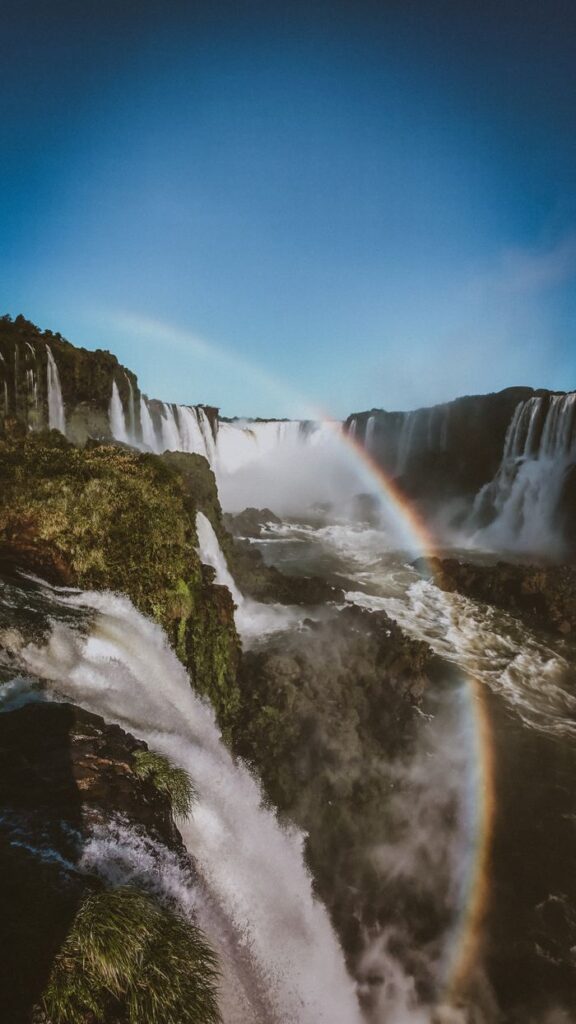 Breathtaking view of Iguazu Falls with a vibrant rainbow under clear blue skies in Brazil.